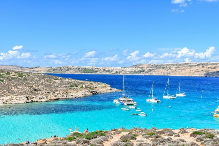 Scenic Blue Lagoon in Comino, Malta, with clear turquoise waters, yachts, and rocky coastline under a bright blue sky.